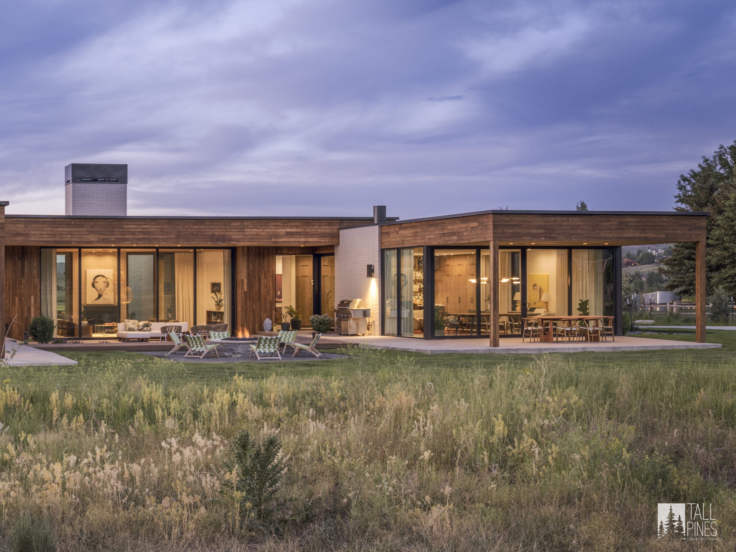 A modern mountain home exterior at dusk with large glass walls and an outdoor seating area, built by Tall Pines Construction in Park City, Utah.