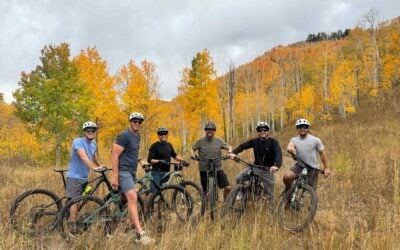 The Tall Pines Construction team stands with their mountain bikes in an open Park City meadow surrounded by fall foliage, reflecting the outdoor lifestyle that inspires their work and community connection.