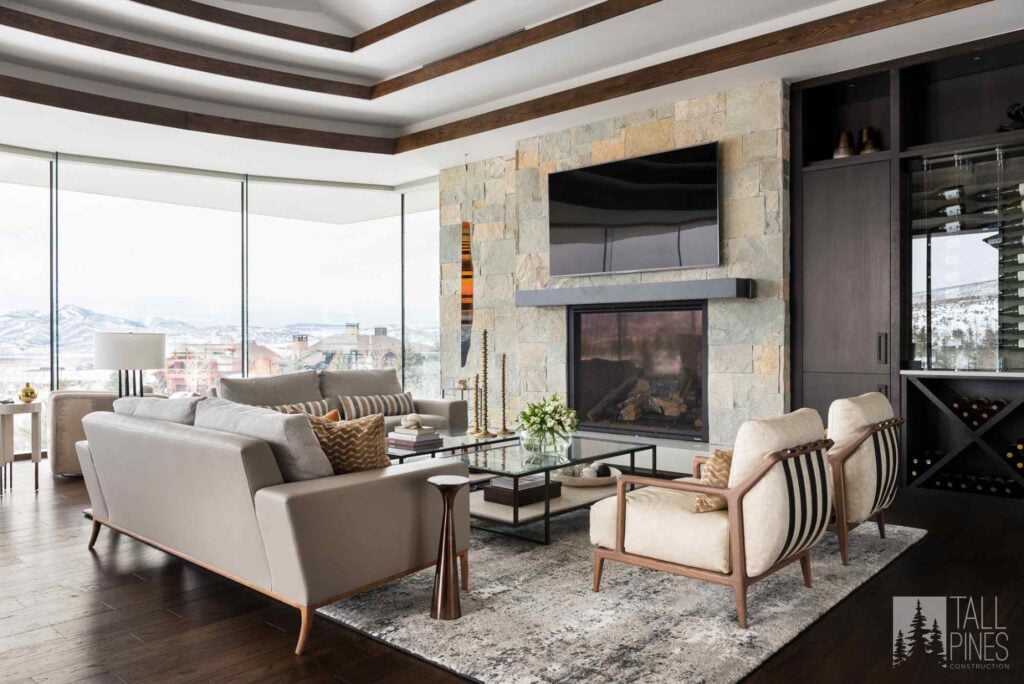 Luxury mountain living room with a stone fireplace, exposed wood beams, floor-to-ceiling windows, and custom finishes.