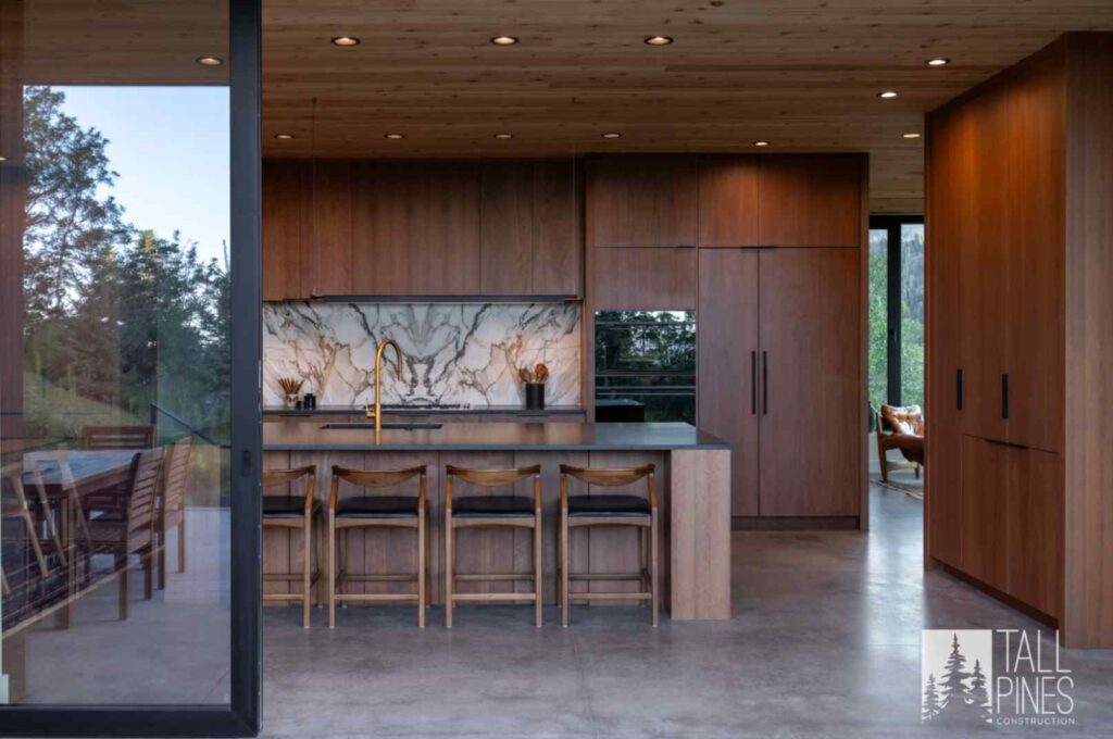 Modern custom home kitchen with warm wood cabinetry, stone backsplash, large island, and floor-to-ceiling windows.