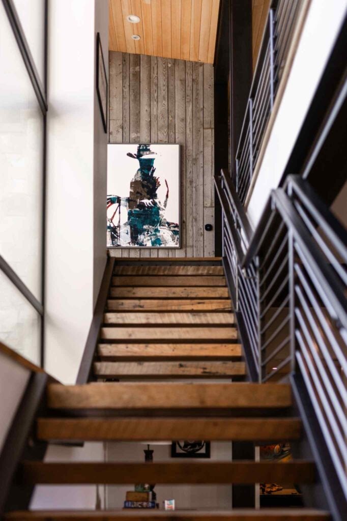 Custom mountain home staircase combining wood treads, black metal railings, reclaimed wood accents, and contemporary artwork.