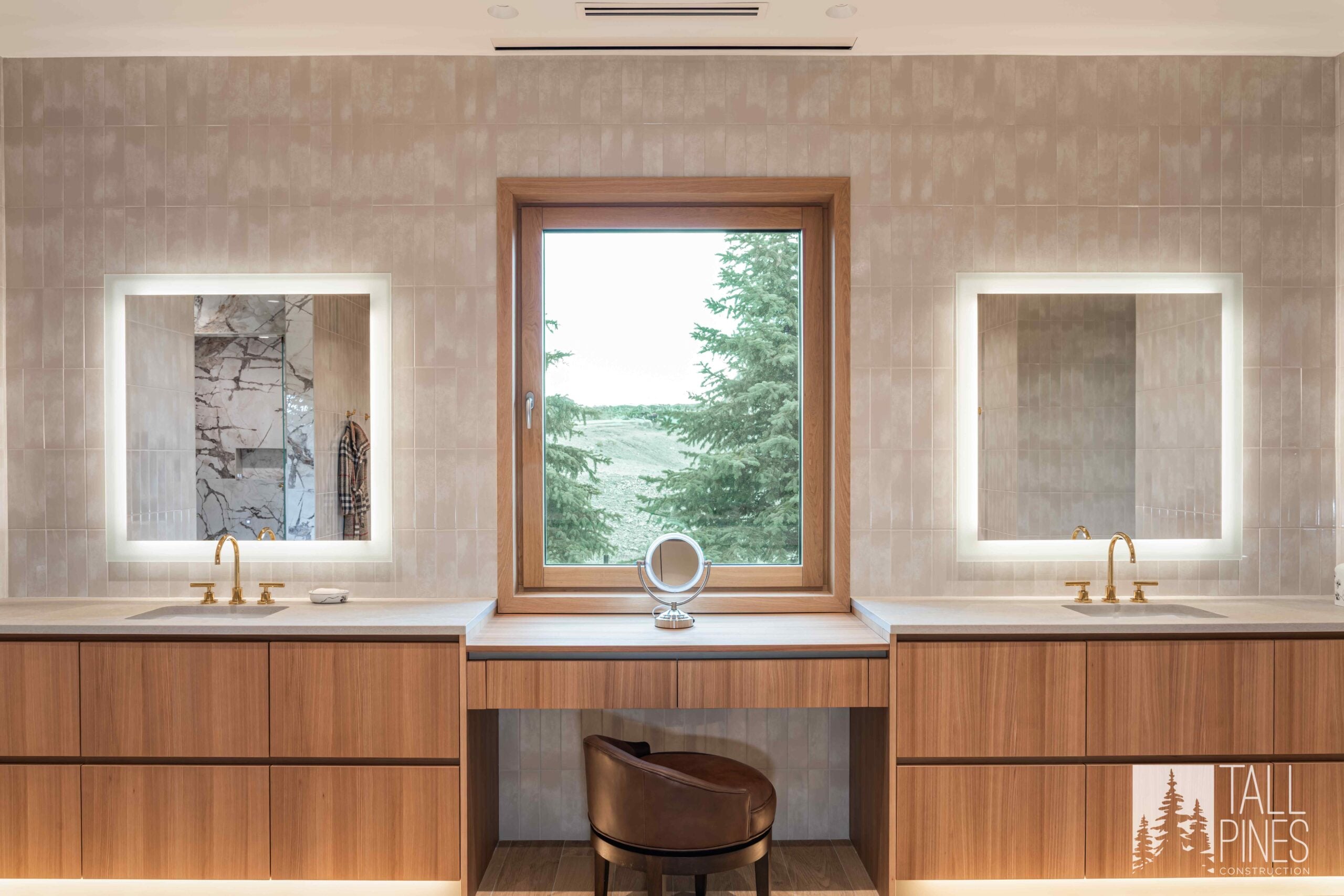 Symmetrical double vanity in the Ridgeline Light bathroom with wood cabinetry, illuminated mirrors, brass fixtures and a central window.